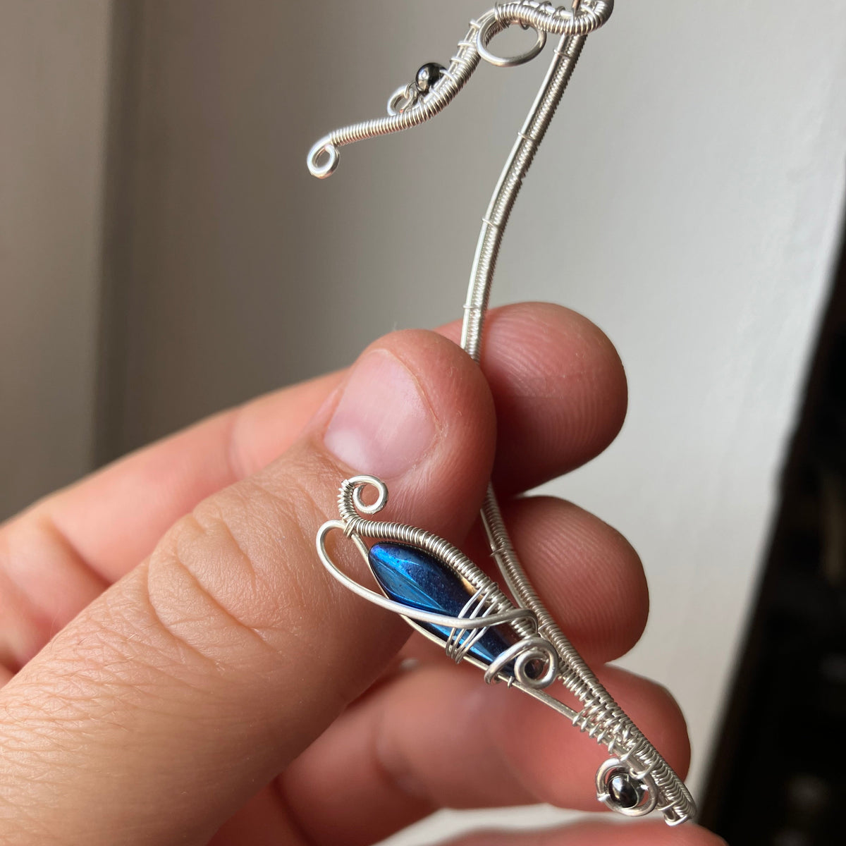 A hand holding a silver wire-wrapped no piercing ear wrap with metallic blue glass bead