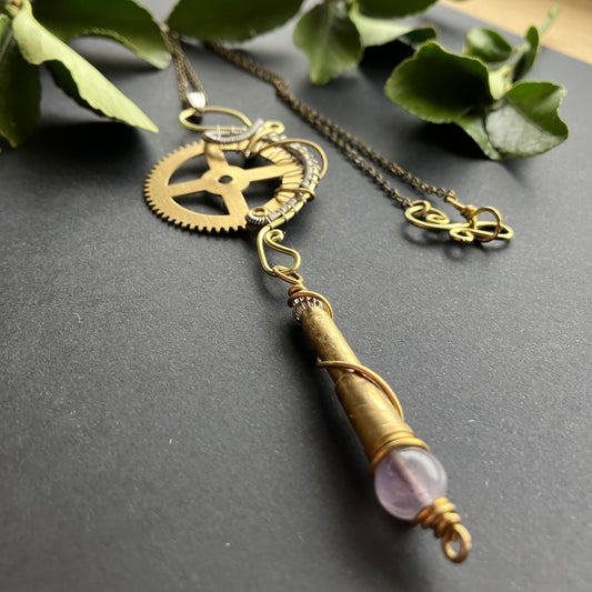 Steampunk-style necklace with gears and a gemstone on a dark surface with green leaves.