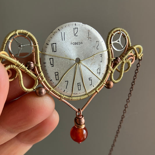 Close-up of a decorative pocket watch with intricate wirework and a red gemstone on a gray background.