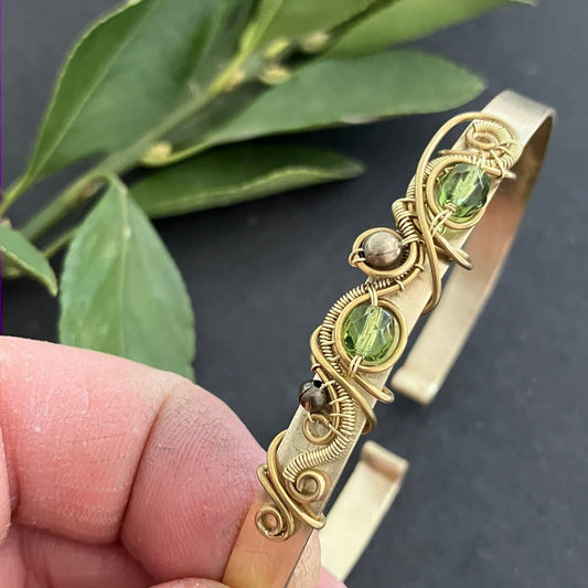 Gold bracelet with green stones held by a hand against a dark background with green leaves.