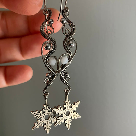 Silver snowflake earrings held by a hand against a gray background